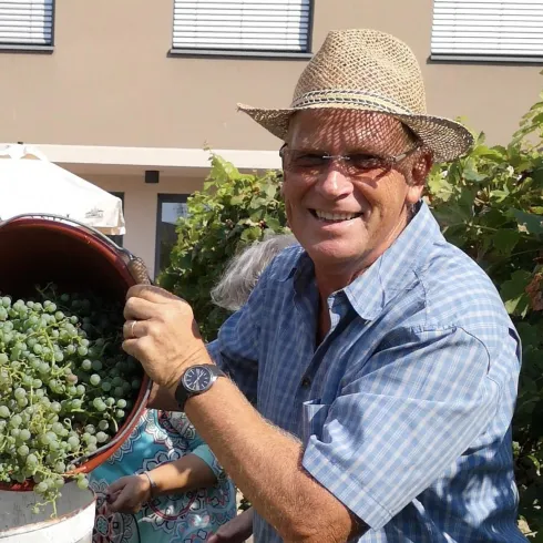 Ein Mann mit einem Hut lächelt und hält einen Eimer mit Trauben. Im Hintergrund sind Weinreben und eine Sonnenterrasse sichtbar.