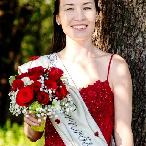 Eine Frau in einem roten Kleid steht neben einem Baum und hält einen Blumenstrauß mit roten Rosen. Sie trägt eine Tiara und ein Schild mit der Aufschrift "Die Schönsten".