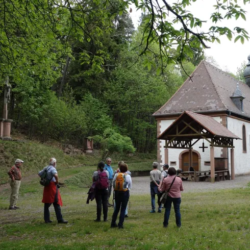 Eine Gruppe von Menschen steht vor einem kleinen, historischen Gebäude im Wald. Umgeben von grünen Bäumen und Natur.