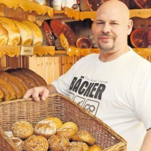 Ein freundlicher Bäcker steht vor einem Regal mit frischem Brot. Er hält einen Korb mit verschiedenen Brötchen.