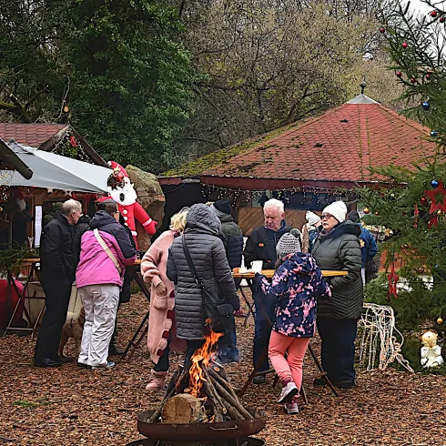 Eine festliche Weihnachtsmarkt-Szene mit Menschen, die um Stände und einen Weihnachtsbaum versammelt sind. Ein Lagerfeuer sorgt für eine gemütliche Atmosphäre.