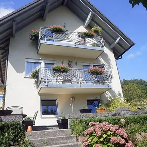 Ein zweistöckiges Haus mit Balkonen, die mit Pflanzen dekoriert sind. Der Himmel ist klar und blau, umgeben von einem grünen Garten.