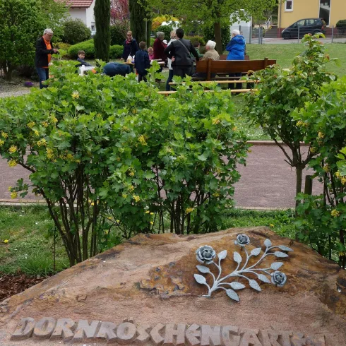 Ein schöner Garten mit einer Steintafel, auf der "Dornröschengarten" steht. Im Hintergrund sind Menschen, die sich versammelt haben.