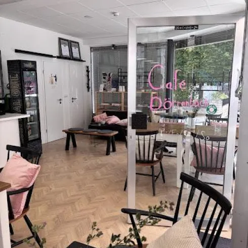 Ein gemütliches Café mit modernen Möbeln und einladenden Dekorationen. Die Fenster ermöglichen viel Tageslicht und eine angenehme Atmosphäre.
