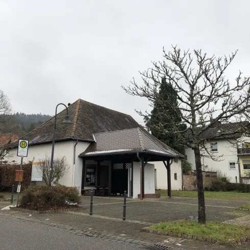 Ein kleiner, weißer Gebäudekomplex mit einem spitzen Dach und einem angrenzenden Parkplatz. Im Hintergrund sind Bäume und ein weiteres Gebäude sichtbar.