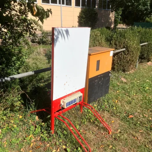 Ein rotes Gestell mit einer weißen Tafel und einem orangefarbenen Kasten steht auf einer Wiese. Im Hintergrund sind Bäume und ein Gebäude sichtbar.
