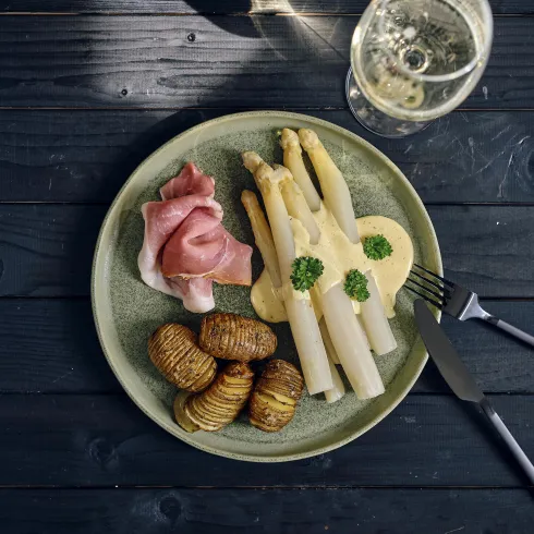 Ein schön angerichteter Teller mit Spargel, Schinken und spiralförmig geschnittenen Kartoffeln. Im Hintergrund steht ein Glaspfang, das ein Getränk enthält.