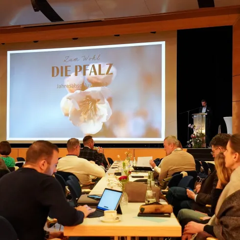 Blick auf eine Bühne mit Präsentation Zum Wohl die Pfalz im Vordergrund Zuschauer