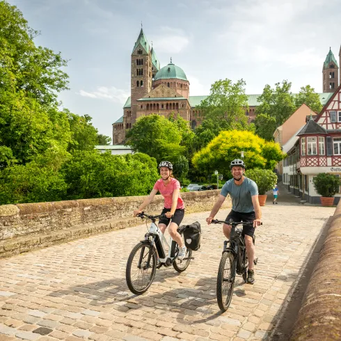 Zwei Personen fahren mit dem Rad über die Sonnenbrücke in Speyer. Im Hintergrund ist der Dom und die Fachwerkhäuser.