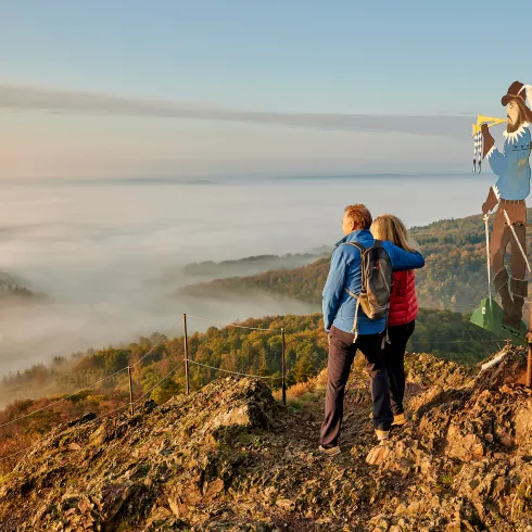 Zwei Personen stehen auf einem Berg im Pfälzer Bergland und blicken auf ein Tal, das von dichtem Nebel bedeckt ist. Neben ihnen ragt die Figur des „Eisernen Mannes“ auf, die majestätisch über der Landschaft thront.
