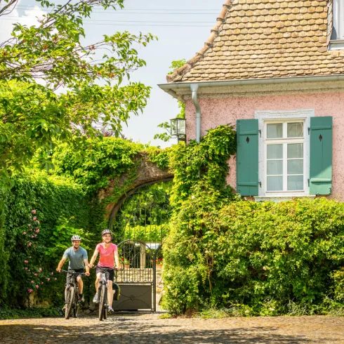 Zwei Personen Fahren mit dem Rad an einem alten rosafarbenen Haus vorbei.