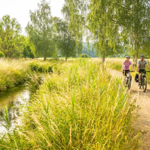 Zwei Personen radeln auf einem Radweg der entlang eines Baches führt.