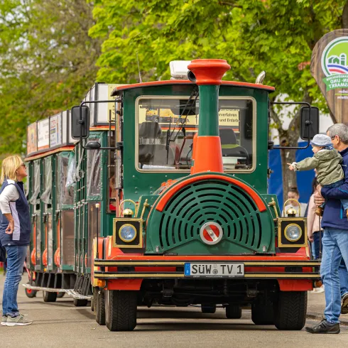 Eine Bimmelbahn schlängelt sich entlang einer Straße. Besuchende eines Marktes bestaunen sie.