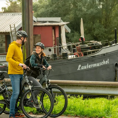 Zwei Personen stehen mit ihrem Rad vor einem Boot mit dem Namen Lautermuschel.