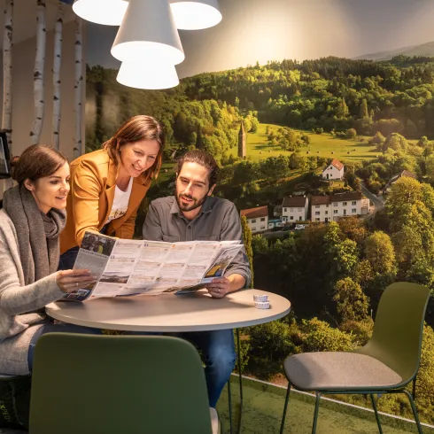 Eine Tourist-Information Mitarbeiterin zeigt zwei Gästen auf einer Pfalzkarte etwas. 