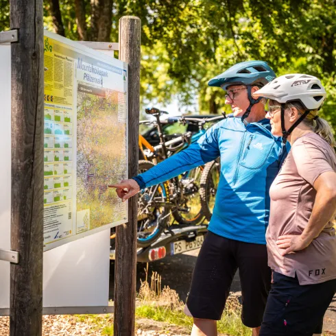 Zwei Personen befinden sich mit ihrem Mountainbike am Haus der Nachhaltigkeit in Johanniskreuz und schauen sich eine Wegekarte an.