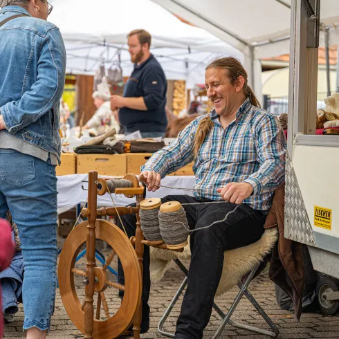 Besuchende schauen einem Marktverkäufer auf dem Keschdemarkt in Hauenstein bei der Arbeit mit der Wollspindel zu.  