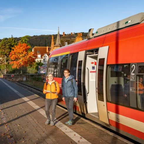 Zwei Personen steigen aus einem Zug. Sie tragen Wanderkleidungen und machen sich auf den Weg zu einer Wanderung in der Pfalz.