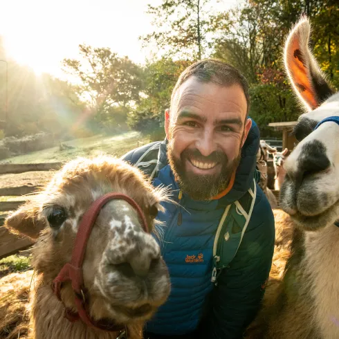 Eine Person lächelt in eine Kamera. Neben der Person stehen zwei Lamas.